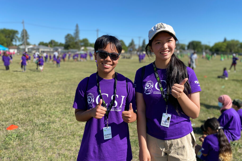 Dean, 14, and his friend Alanna, 13, smile brightly as they stand happily in an open field at the Jumpstart Games, with kids running energetically behind them.
